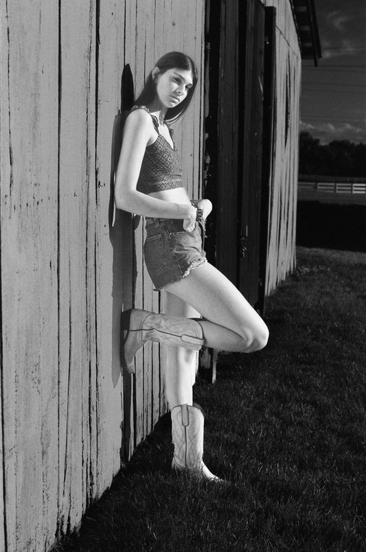 Black-and-white portrait of a young woman with dramatic side lighting, emphasizing the contours of her face and body leaning against the barn