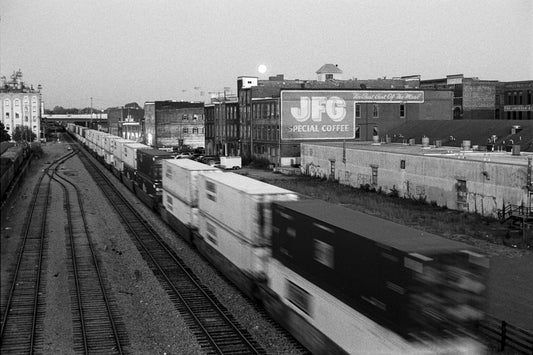 Black and white photograph of a freight train moving along tracks in an industrial city setting, with buildings and a large JFG Special Coffee sign visible in the background.