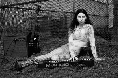 Black-and-white portrait of a young woman playing keyboards plugged into an amp outdoors with a guitar in the background
